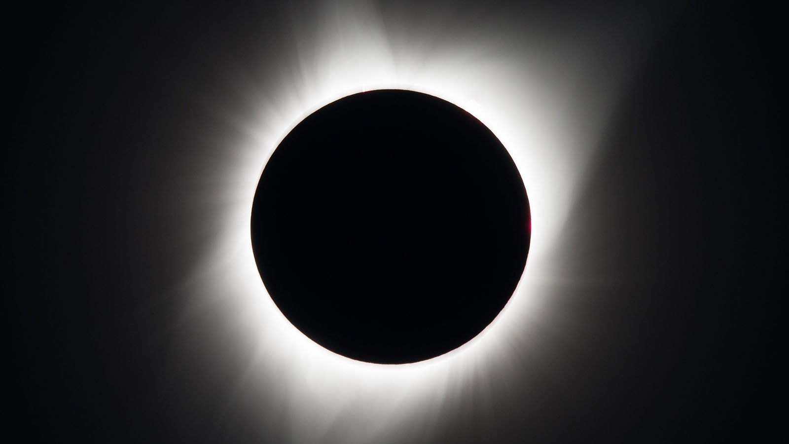 A total solar eclipse is shown as light rays emit from a black circle against a black sky.